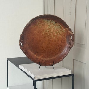 Könnte beinhalten: Ein großes, rundes, braunes Serviertablett aus Keramik mit zwei Griffen. Das Tablett hat eine gesprenkelte, rustikale Oberfläche und wird auf einem schwarzen Metallständer präsentiert.