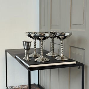 Puede incluir: Conjunto de copas de champán plateadas y una pequeña copa con tallo retorcido. Las copas están dispuestas sobre una estantería negra. El fondo incluye una pared blanca y una puerta gris. Perfectas para brindar.