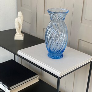 May include: A blue glass vase with a swirl design sits on a white book on a black metal shelf. A white sculpture of two faces is on the shelf behind the vase.