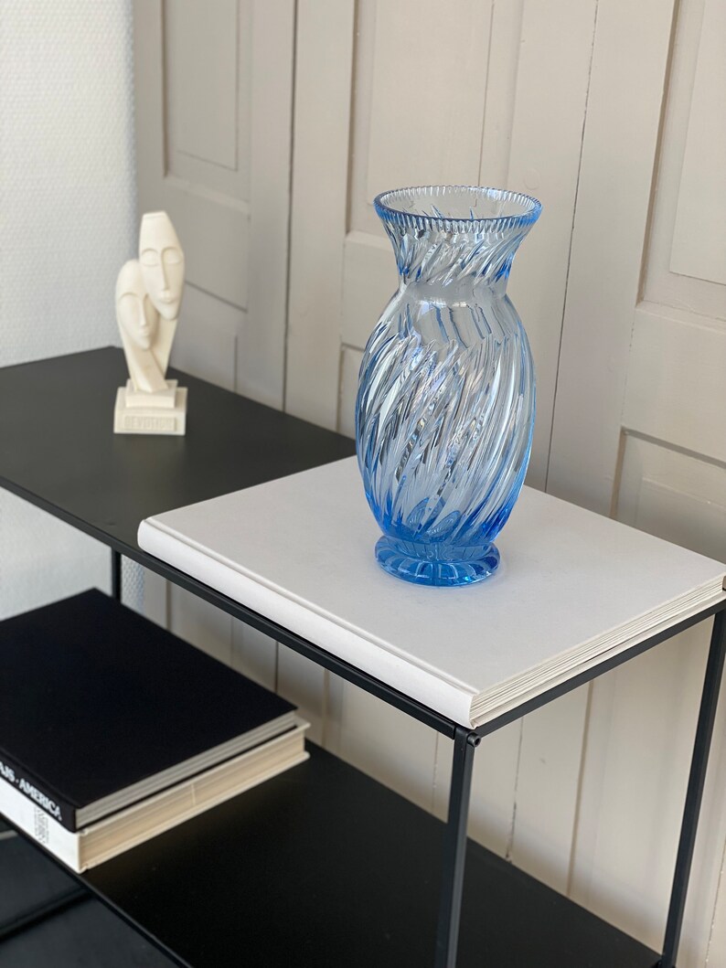 May include: A blue glass vase with a swirl design sits on a white book on a black metal shelf. A white sculpture of two faces is on the shelf behind the vase.