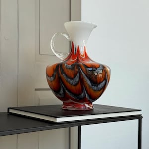 Könnte beinhalten: Eine dekorative Glaskaraffe mit weißem Oberteil und Griff, die in einen orange-schwarz gemusterten Körper übergeht. Die Karaffe steht auf einer schwarzen Oberfläche und zeigt ihr einzigartiges Design und ihre Handwerkskunst.