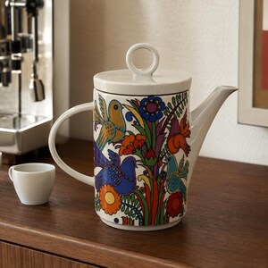 May include: A white ceramic coffee pot featuring a vibrant, multi-coloured floral and bird pattern. The pot has a cylindrical body, a curved handle, a pointed spout, and a flat lid with a circular handle on top. Two small white cups sit nearby.