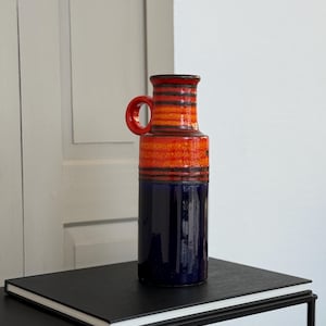 Puede incluir: Un jarrón de cerámica alto con asa, con una llamativa combinación de colores. La sección superior es naranja con rayas negras, que transiciona a una base azul oscuro. El jarrón mide unos 30 cm de alto.