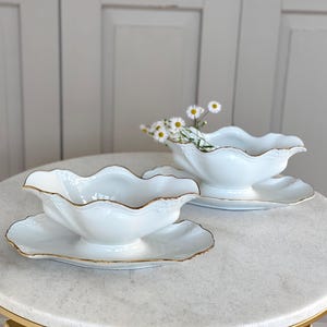 Op de afbeelding: Twee witte porseleinen juskommen met gouden rand en bijpassende borden. De kommen hebben een gegolfde rand en staan op een marmeren tafel met een gouden onderstel.