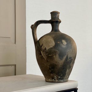 May include: A dark brown ceramic pitcher with a handle and a spout. The pitcher has a rustic, aged look with a white glaze in some areas.