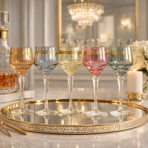 May include: Five colorful wine glasses with gold rims are displayed on a gold-rimmed tray. The glasses are in shades of amber, blue, yellow, pink, and green. A crystal decanter and a bouquet of white roses are in the background.