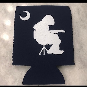 May include: A navy blue can cooler with a white silhouette of a person playing a guitar. The person is sitting on a stool and there is a crescent moon in the upper left corner.