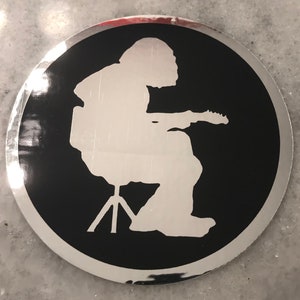 May include: A silver and black round sticker with a silhouette of a person sitting on a stool and playing a guitar.