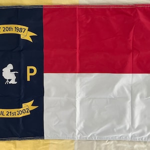 May include: A rectangular flag with a design featuring a navy blue vertical stripe on the left, and a red and white horizontal stripe on the right. The navy stripe has gold text and a silhouette of a person sitting in a chair.