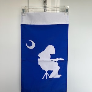 Puede incluir: Una bandera azul y blanca con una silueta blanca de un guitarrista, una luna creciente y un borde superior blanco. La bandera está colgada de una percha de plástico transparente.