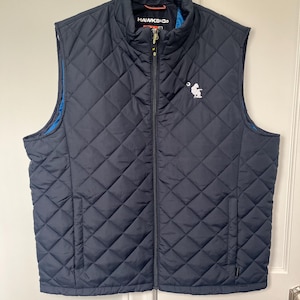 Peut inclure: Une veste matelassée bleu marine avec une fermeture éclair et un petit logo blanc sur la poitrine. La veste a un motif en losange et une doublure bleue.