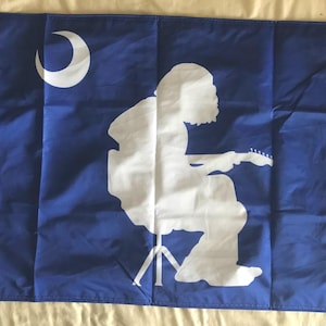 May include: A blue flag featuring a white silhouette of a person playing a guitar, seated on a stool. A white crescent moon is in the upper left corner. The flag has a white border with grommets.