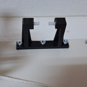 Può includere: Primo piano di un pezzo nero stampato in 3D con due pezzi di plastica bianca inseriti nella parte superiore. Il pezzo è fissato a una superficie bianca con quattro viti.