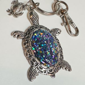 May include: A silver turtle keychain with a glittery purple, blue, and green shell. A small silver crab charm is attached to the keychain ring.