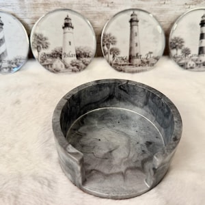 May include: Four circular coasters featuring black and white lighthouse illustrations with text labels St. Augustine, FL, Amelia Island, FL, Ponce Inlet, FL, and Cape Canaveral, FL, accompanied by a matching gray and white marbled resin coaster holder.