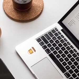 May include: A silver laptop computer with a black keyboard and a black trackpad. A brown coffee cup sticker with a smiley face is on the laptop.