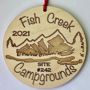 Puede incluir: Un adorno redondo de madera con el texto "Fish Creek Campgrounds" y "2021" grabado en él. El adorno también presenta una imagen estilizada de montañas y árboles, y el texto "SITE #242".