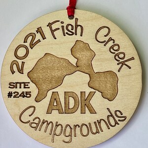 Puede incluir: Un adorno de madera con el texto "2021 Fish Creek ADK Campgrounds" y "Site #245". El adorno presenta una silueta de un lago.