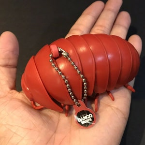 Può includere: Un millepiedi giocattolo di plastica rosso segmentato con una catena argentata e un'etichetta rosa che dice "Bango Brush".