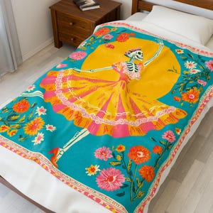 May include: A turquoise blanket featuring a skeleton in a yellow dress dancing in front of a yellow sun. The blanket is decorated with pink, orange, and white flowers. The edges have a decorative border.