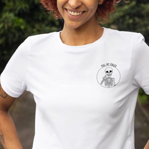 Puede incluir: Camiseta blanca con un gráfico en blanco y negro de un esqueleto dentro de un círculo, con el texto "PULL MY FINGER". El esqueleto está haciendo una señal de pulgar hacia arriba. La camiseta está hecha de un material suave.