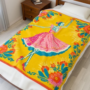May include: A yellow blanket featuring a dancing skeleton in a pink polka-dot dress and blue accents. The blanket is decorated with a floral border in orange, pink, and teal. The design is reminiscent of Day of the Dead celebrations.