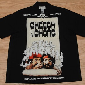 May include: Black short-sleeved button-up shirt with a white print of Cheech & Chong from the film "Up in Smoke". The print features the text "Still Smokin'" and the tagline "They'll have you rollin' in your seats!"