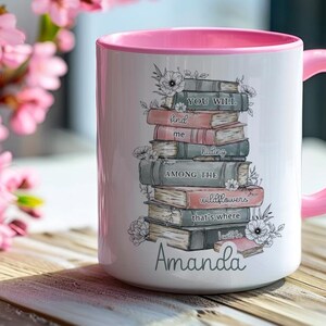 Op de afbeelding: Witte keramische mok met een roze handvat en rand, met een stapel boeken met bloemaccenten en de tekst "You will find me among the wildflowers that's where I'll be." De naam "Amanda" staat onderaan.
