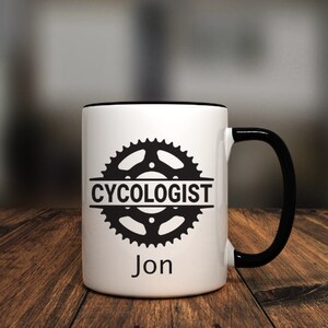 May include: White ceramic mug with a black rim and handle. The mug features a black and white graphic of a bicycle chainring with the text "CYCOLOGIST" and the name "Jon" printed below.