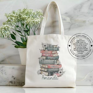 May include: Cream-colored tote bag featuring a stack of books with floral accents. The books are in various colors, with titles visible. The name "Amanda" is written below the books. A stamp on the bag reads "100% Hand Drawn Unique Artwork."