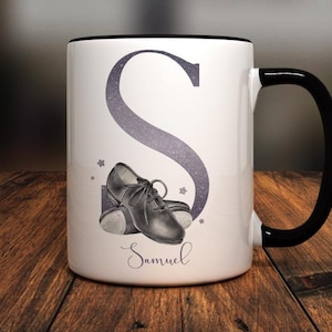 Könnte beinhalten: Eine weiße Keramiktasse mit schwarzem Henkel. Die Tasse zeigt einen großen, glitzernden lila Buchstaben "S" und ein Paar schwarze Steppschuhe. Der Name "Samuel" ist unter den Schuhen aufgedruckt.