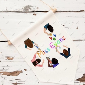 May include: A white banner with a wooden dowel and string for hanging. The banner features a colorful illustration of children and the text "Miss Evans". The children are depicted with art supplies and a rainbow.