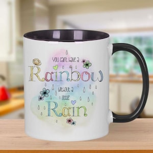 Może przedstawiać: Biały ceramiczny kubek z czarnym wnętrzem i uchwytem. Kubek zawiera napis "You can't have a Rainbow without a little Rain" w kolorowej, fantazyjnej czcionce, z ilustracjami kwiatów i kropli deszczu.