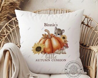 May include: White square cushion featuring a watercolor illustration of a mouse, pumpkin, sunflowers, and mushrooms. Text on the cushion reads "Binnie's little AUTUMN CUSHION". The cushion is placed on a wicker chair.