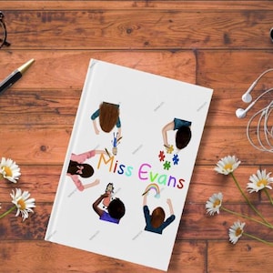 May include: A white notebook with the text "Miss Evans" and illustrations of children, surrounded by a pen, glasses, a smartphone, headphones, a cup of coffee, and daisies on a wooden surface.