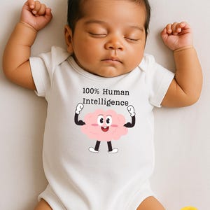 May include: A sleeping baby wearing a white onesie with the text "100% Human Intelligence" and a cartoon brain graphic. The baby has dark hair and is lying on a light-colored surface.