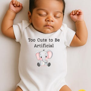 May include: A white baby onesie with the text "Too Cute to Be Artificial" and a cartoon elephant illustration. The baby is lying down with their eyes closed.