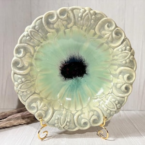 May include: A decorative ceramic plate with a raised floral design in a light green and white color scheme. The center of the plate features a stylized flower with a black and blue center.