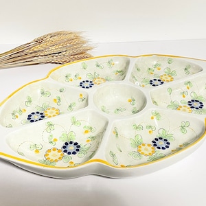 May include: A white ceramic serving dish with six individual compartments. The dish is decorated with a floral pattern in shades of yellow, green, and blue.