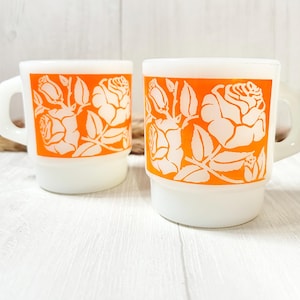 May include: Two white ceramic mugs with orange floral designs. The mugs have a simple, classic shape and are perfect for everyday use.