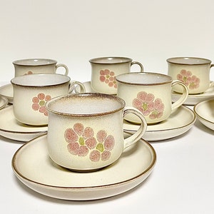 May include: Set of eight cream-coloured teacups and saucers with a floral design. The cups and saucers feature a brown rim and a pink and green floral pattern.