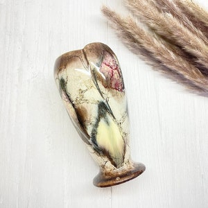 May include: A ceramic vase with a distinctive, organic form. The vase displays a mix of brown, cream, and beige hues with a crackle glaze. The base is a darker brown, and the vase has a flared, rounded top. Dried ornamental grass is in the background.