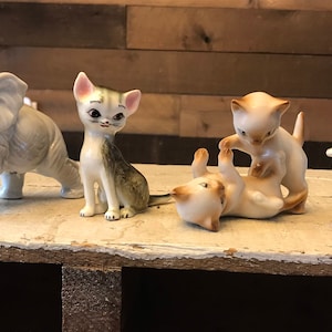 May include: A collection of five porcelain figurines, including a grey elephant and four cats. The cats are in various poses, including one cat sitting up, one cat lying down, and two cats playing. The figurines are all hand-painted and have a vintage look.