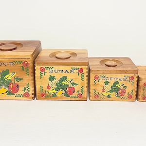 May include: Set of four wooden canisters with lids. Each canister has a different fruit design and label: Flour, Sugar, Coffee, and Tea.