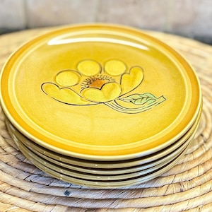 May include: A stack of six yellow ceramic plates with a floral design. Each plate features a yellow flower with black outlines and a green leaf. The plates have a yellow border and are stacked on a woven mat.