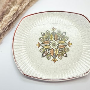 May include: A white ceramic serving platter with a brown and green floral design. The platter has a raised rim and a speckled finish.