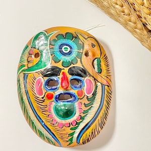 May include: A hand-painted ceramic mask with a colorful design featuring a face with a large nose, eyes, and mouth, surrounded by flowers and birds. The mask is painted in shades of yellow, green, blue, red, pink, and black.