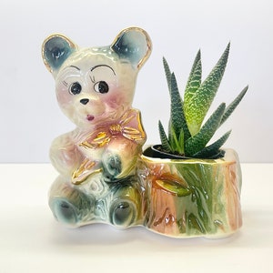 May include: A ceramic planter shaped like a teddy bear holding a bow. The planter is painted in shades of pink, blue, and green. A small succulent plant is growing in the planter.