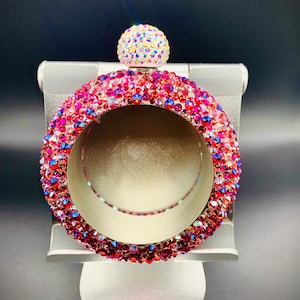 May include: A pink and red crystal-encrusted bangle bracelet with a clear crystal ball on top. The bracelet is on a silver stand.