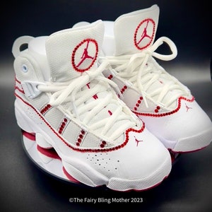 May include: White leather trainers with red accents and rhinestones. The trainers have a red Jumpman logo on the side and a red circle with a Jumpman logo on the tongue.